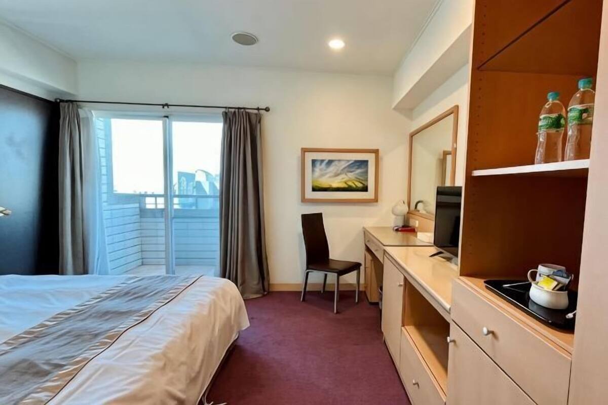 A hotel room with a bed, desk, chair, and a view of the city through the window.