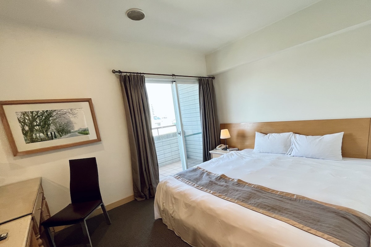 A hotel room with a large bed, a desk with a chair, a framed picture on the wall, and a window with curtains.