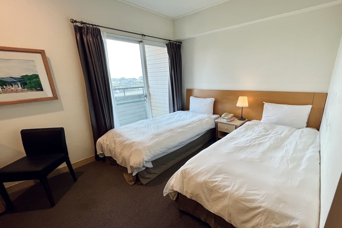 A hotel room with two beds, a desk, a chair, and a window with curtains.