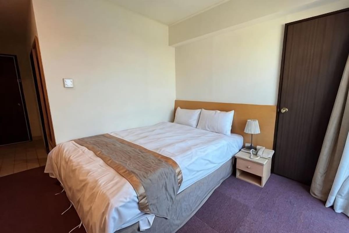 A hotel room with a bed, bedside table, lamp, and a door.