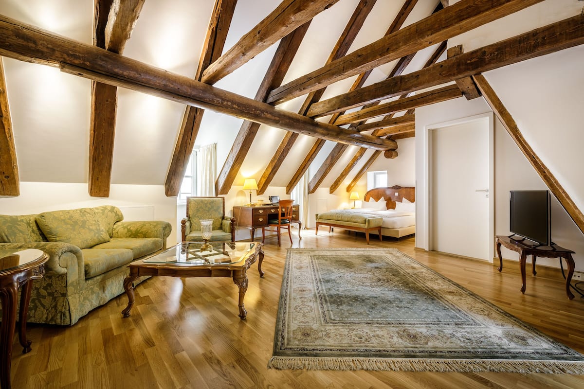 Un loft espaiós amb bigues de fusta al descobert, un sofà verd, una taula de cafeïna de vidre, un penjall estampat i un llit amb televisió.