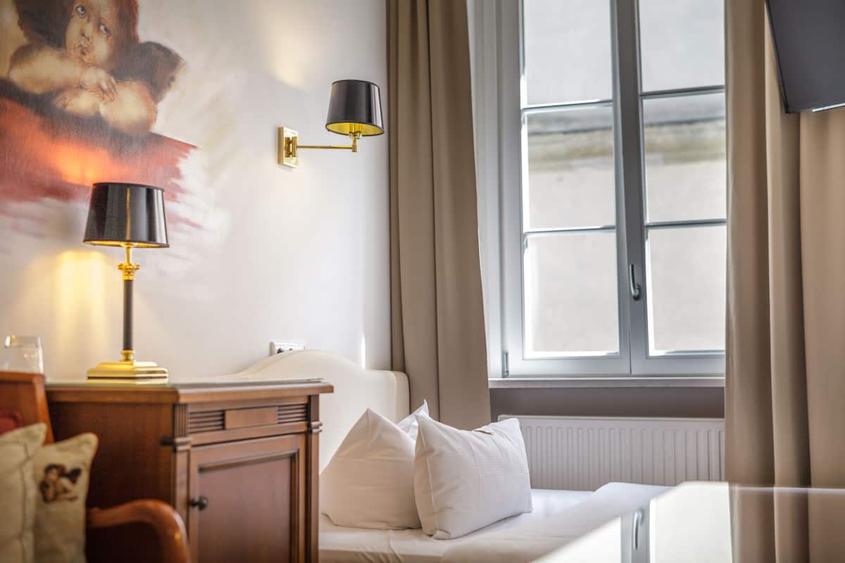 Habitació d'hotel amb llit, calaixera, làmpada, llum penjada a la paret i finestra amb cortines.