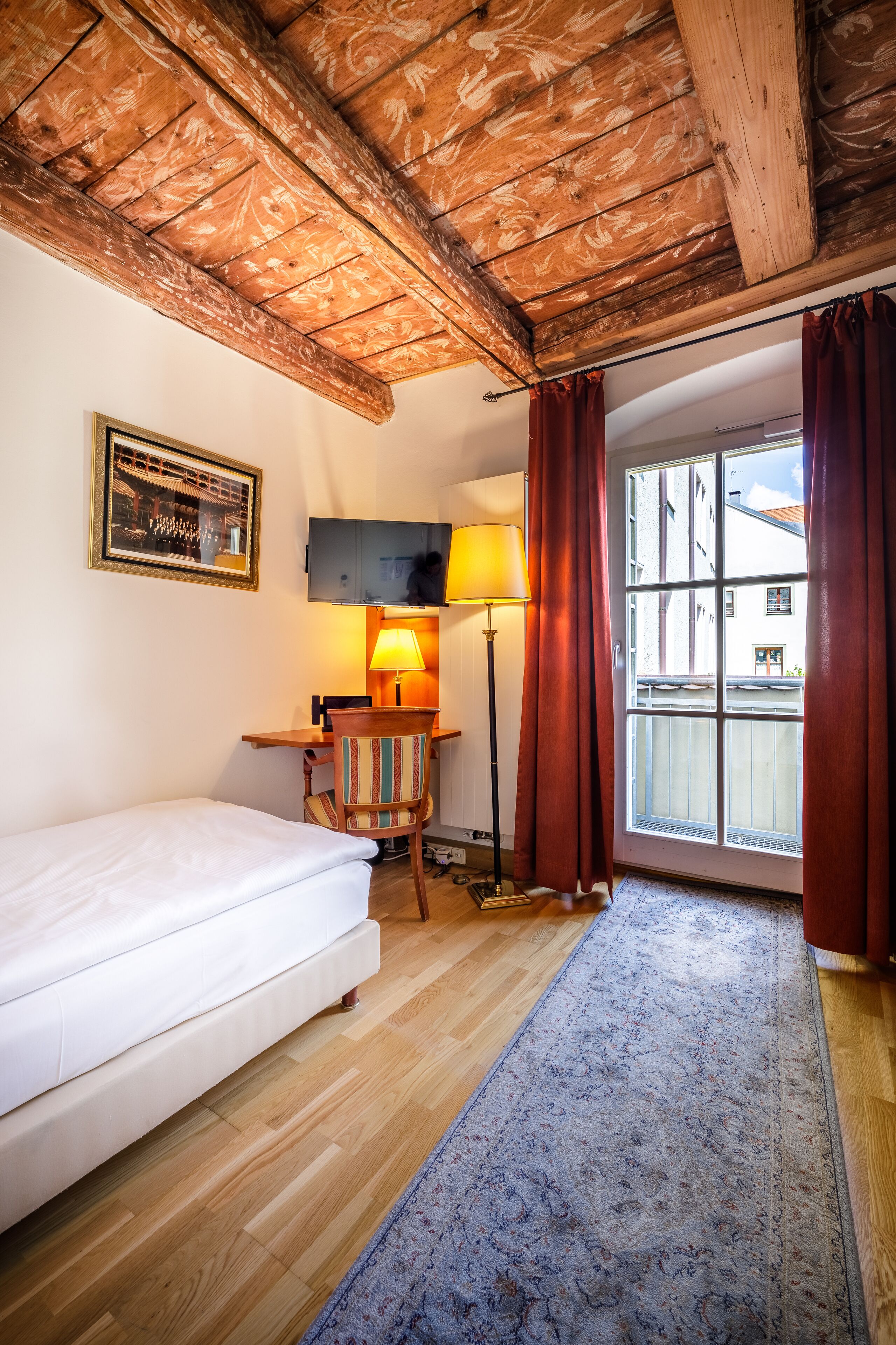 Una habitación con techo de madera, una cama, un escritorio con lámpara, una silla y una ventana con cortinas.