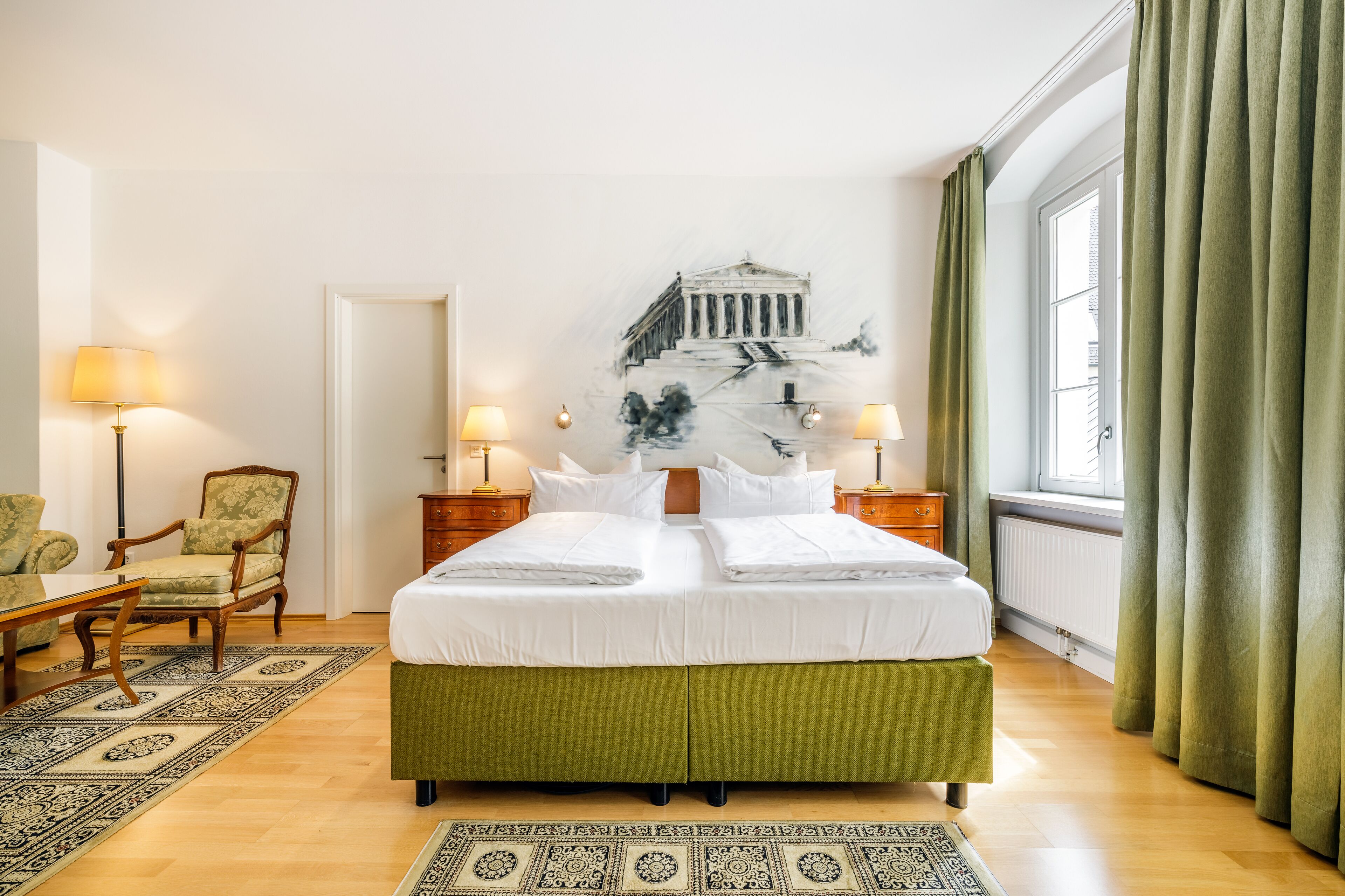 Una habitación de hotel con una cama grande, dos mesitas de noche con lámparas, una silla, un tapete estampado y un ventanal con cortinas.