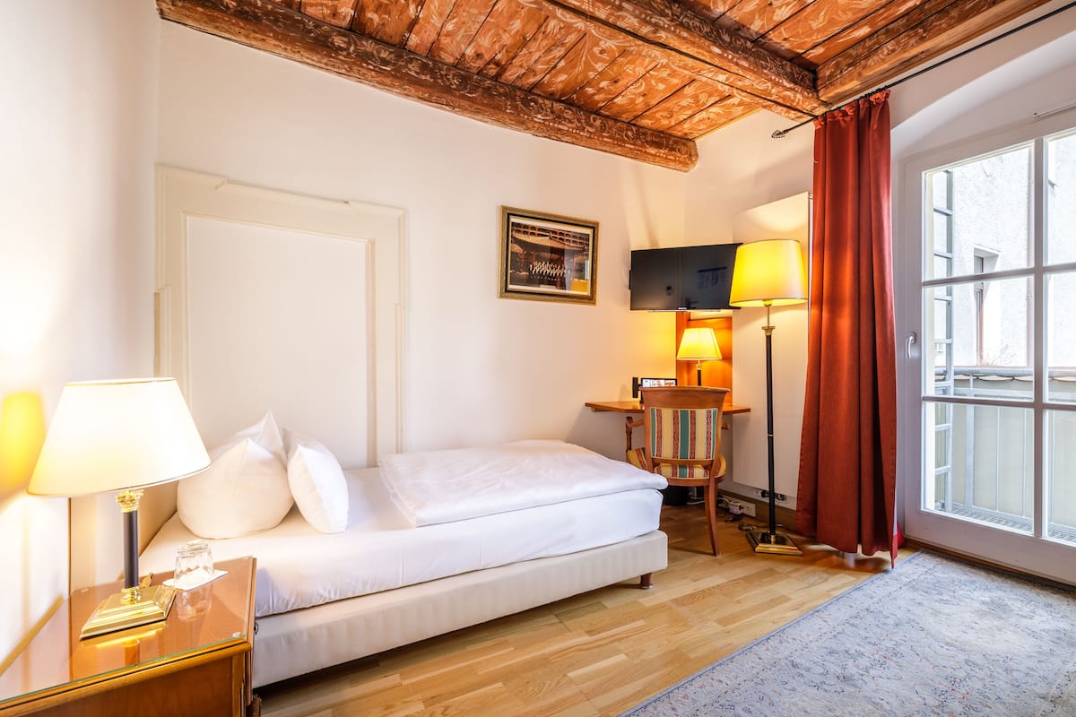 Habitació d'hotel amb llit, una petita taula de nit amb làmpada, escriptori amb cadira, televisió i finestra amb cortines.