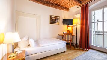 A hotel room with a bed, a nightstand with a lamp, a desk with a chair, a television, and a window with curtains.