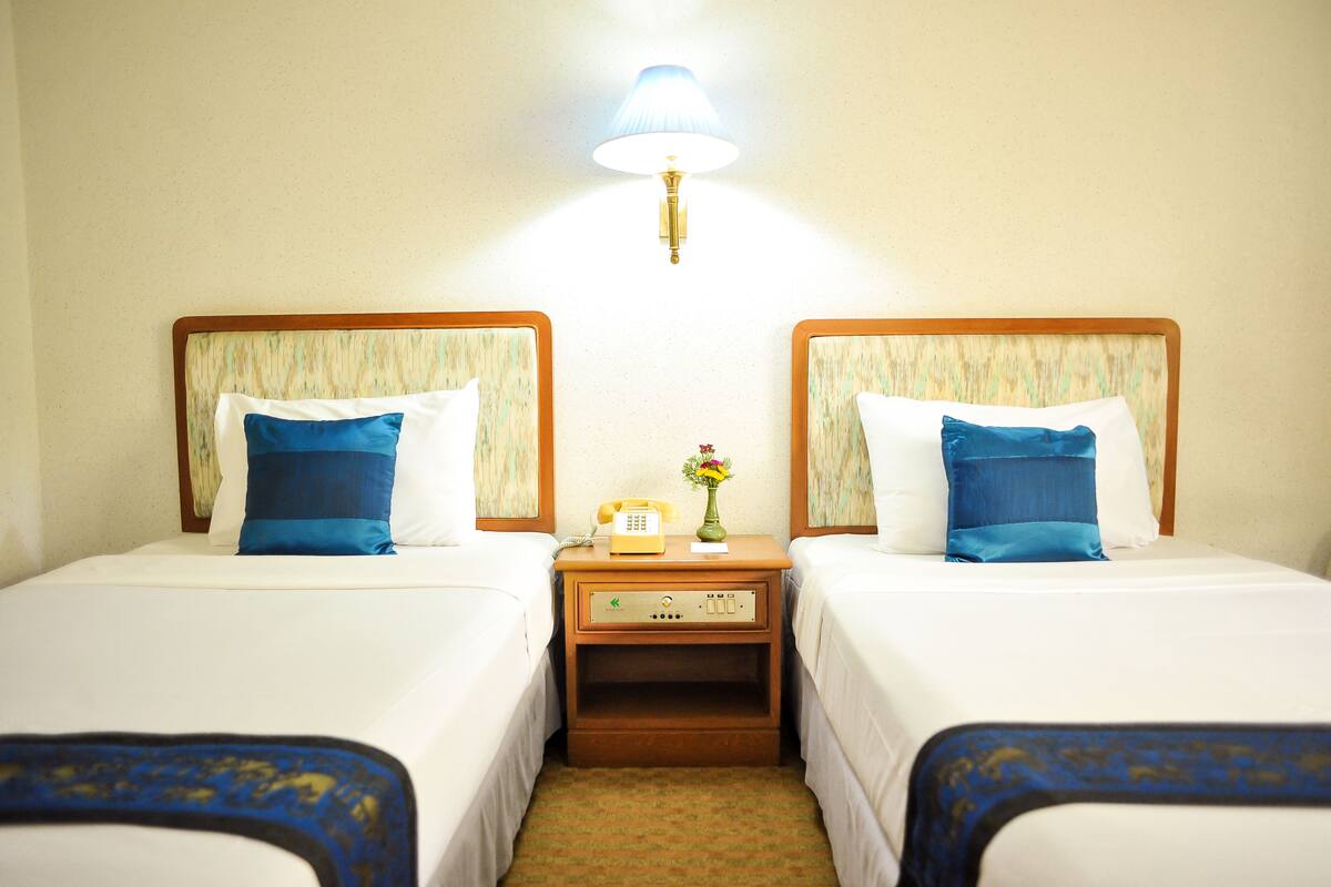 Quarto de hotel com duas camas, cada uma com travesseiro azul, uma mesa de cabeceira com luminária e telefone.