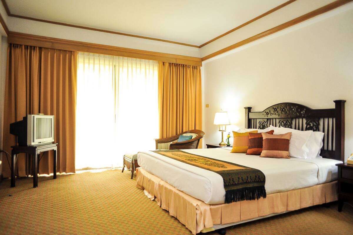 Quarto de hotel com uma cama grande, uma televisão sobre uma mesinha, um abajur e uma cadeira.