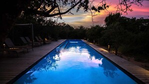 Piscina con sillones de descanso y muelle de madera, rodeada de árboles y un cielo al atardecer.