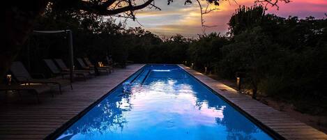 A swimming pool with lounge chairs and a wooden deck, surrounded by trees and a sunset sky.