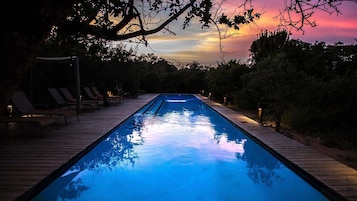 Piscina com espreguiçadeiras e deck de madeira, cercada por árvores e céu ao pôr do sol.