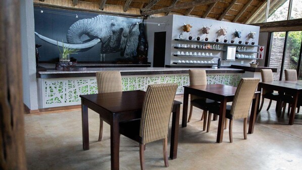 A restaurant with a large elephant mural, a bar area with shelves of bottles and glasses, and a seating area with striped chairs.
