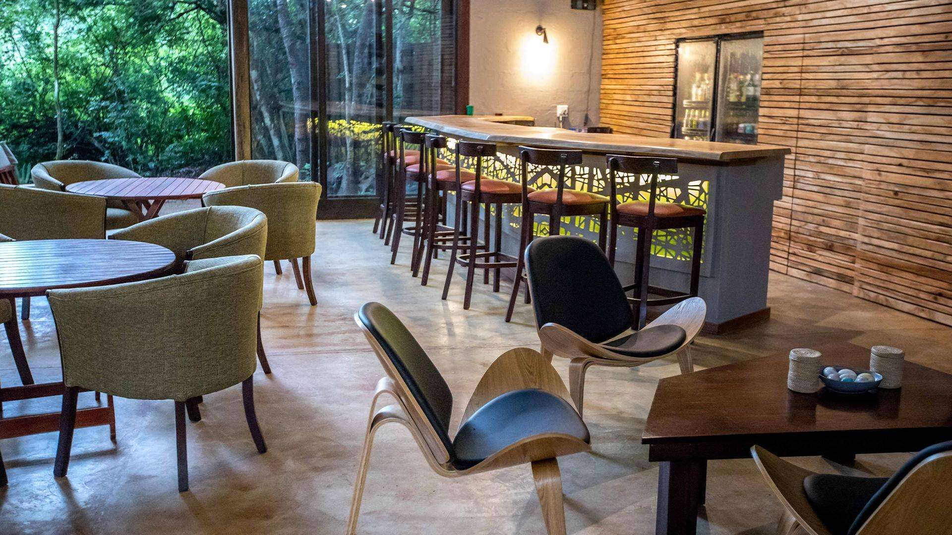 Un comedor moderno con mesas y sillas de madera, una zona de bar con taburetes y ventanas amplias que ofrecen vistas de la vegetación circundante.