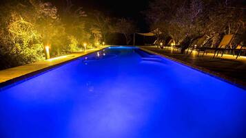 Una piscina iluminada con luces azules durante la noche, rodeada de árboles y mobiliario exterior.