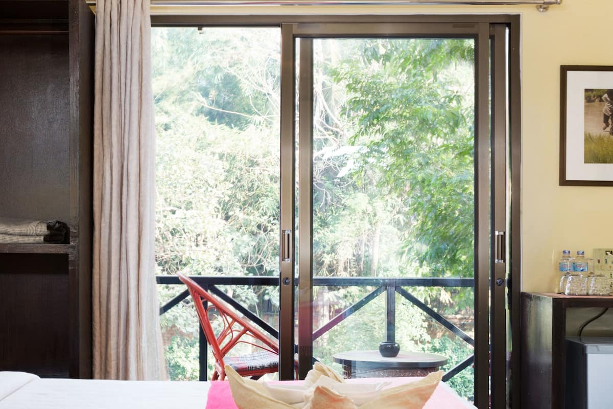 Quarto de hotel com cama, colcha vermelha, janela com vista para árvores, varanda com cadeira e uma mesa pequena.