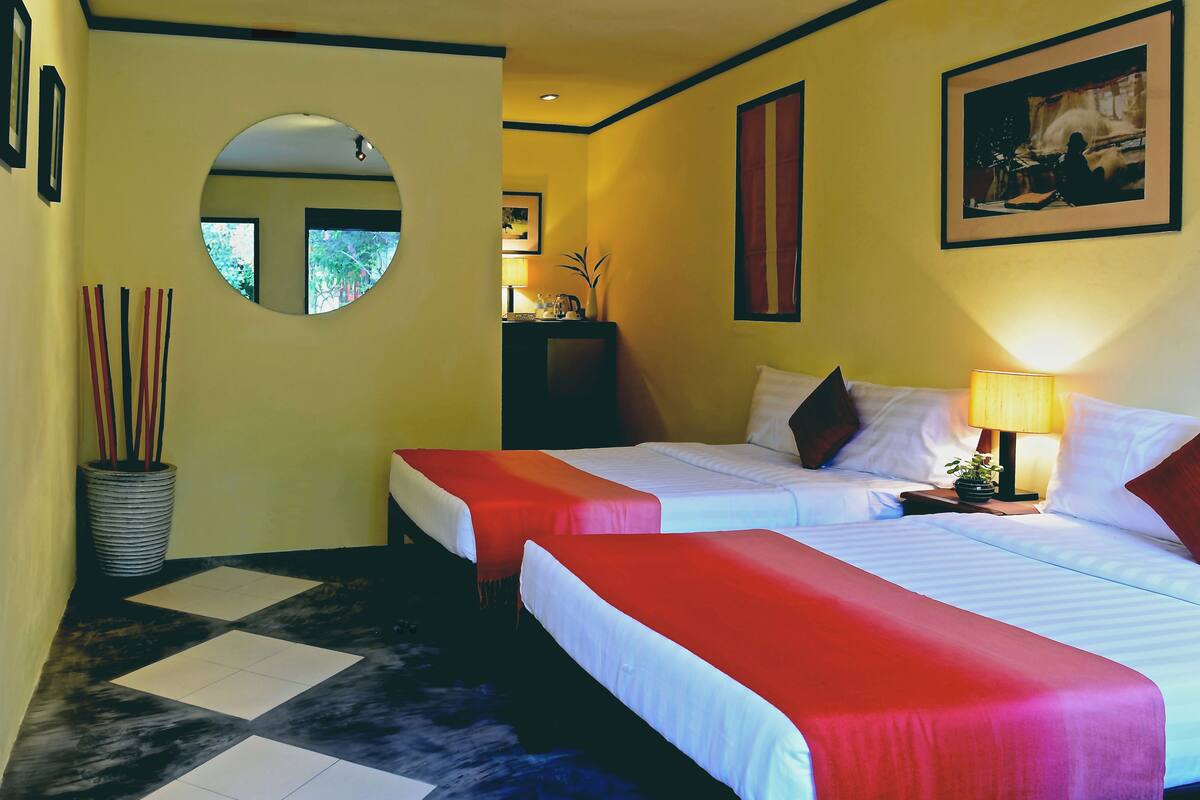 Quarto de hotel com duas camas, um espelho redondo, uma pequena mesa com abajur e um vaso de planta.