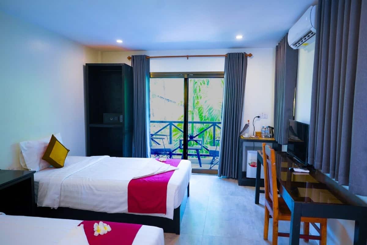 Quarto de hotel com duas camas, uma mesa com cadeira, televisão e varanda com vista para área verde.
