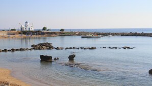 Escena costera con un edificio blanco de cúpulas azules, una playa arenosa y agua tranquila con rocas.
