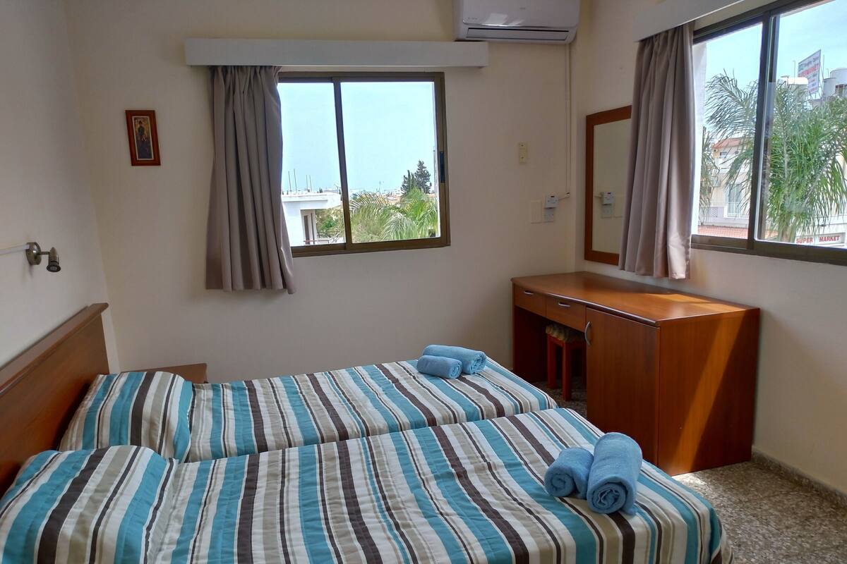 A hotel room with two beds, a wooden dresser, a window with curtains, and a view of palm trees and buildings.