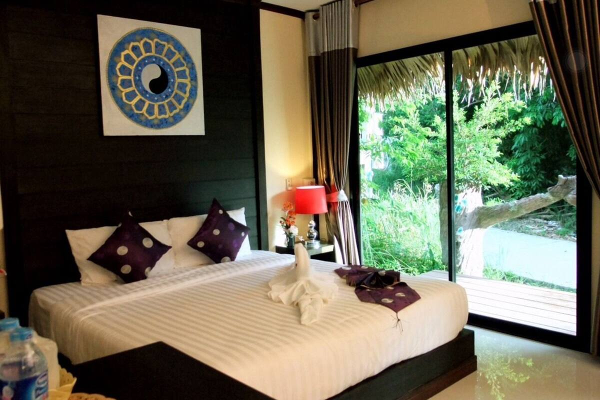 A hotel room with a large bed, a wall-mounted artwork, a window with curtains, and a view of greenery outside.