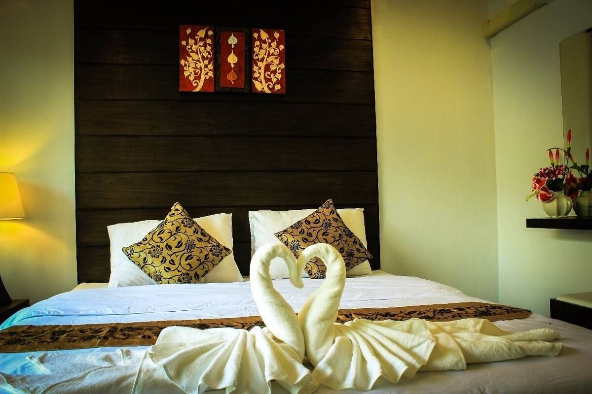 A hotel room with a bed, decorative pillows, a wall with a wooden panel, and a nightstand with a lamp.