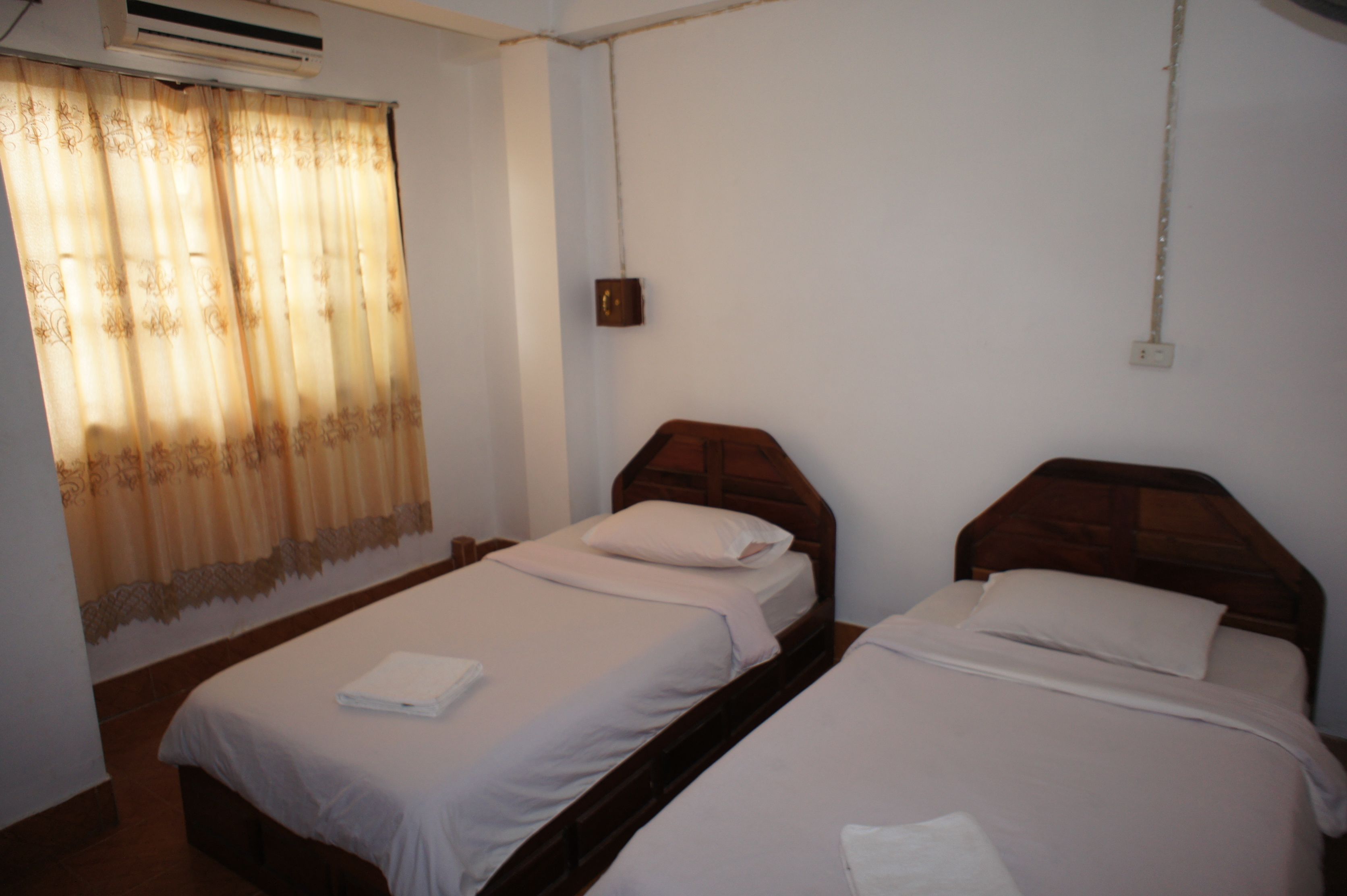 A room with two single beds, a wooden headboard, a curtain, and a wall-mounted air conditioner.
