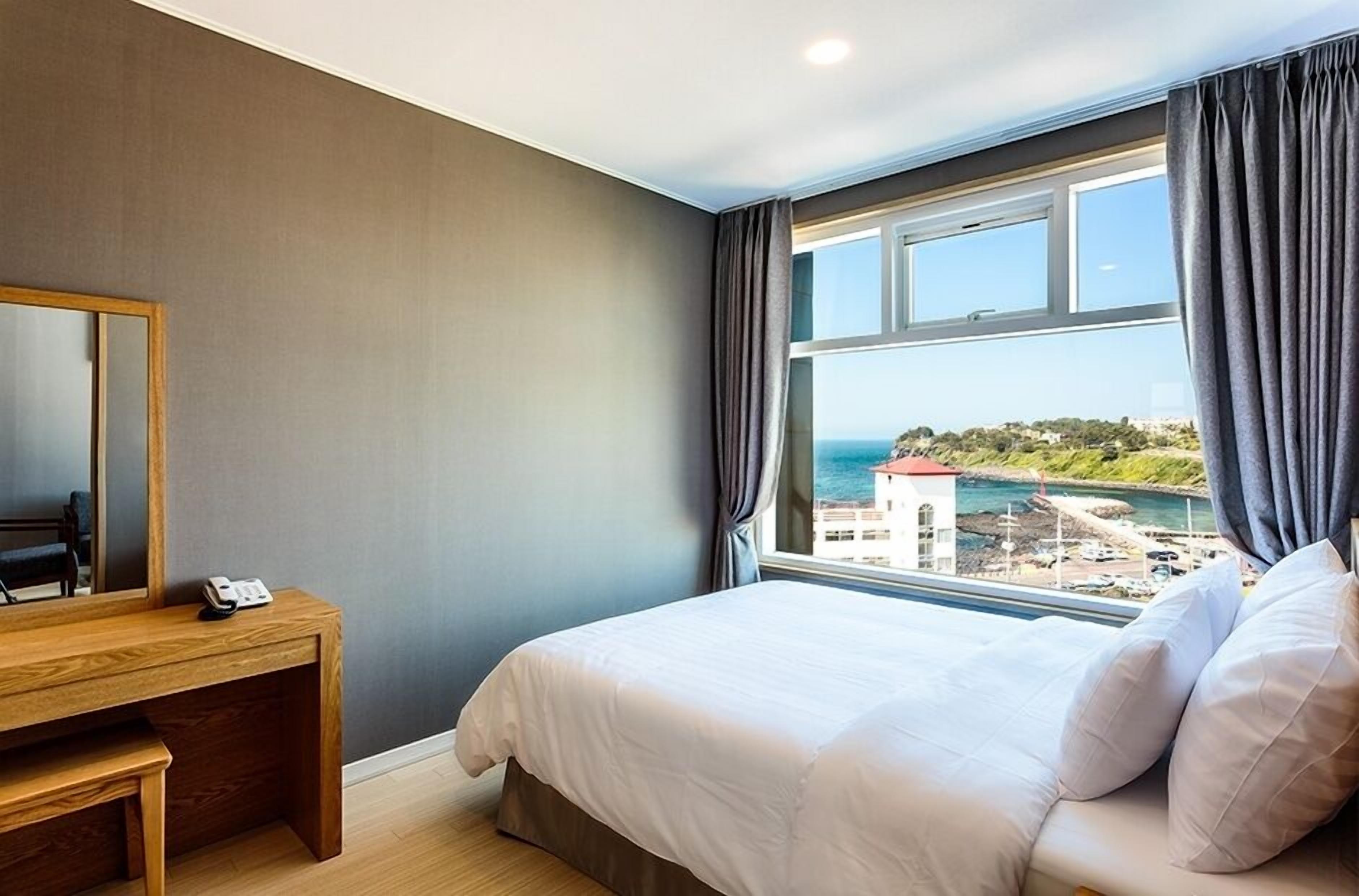 A bedroom with a bed, a wooden desk, a telephone, and a view of the sea.