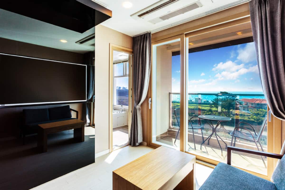Een moderne hotelkamer met een grote televisie, een houten bankje, een klein tafeltje en een balkon met uitzicht op de stad en de oceaan.
