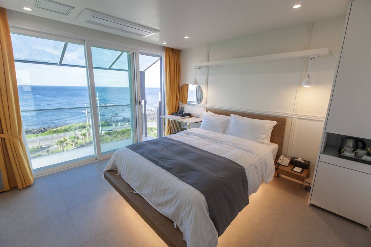 Een moderne hotelkamer met een groot bed, een bureau met telefoon en uitzicht op de oceaan door middel van kamerhoge ramen.