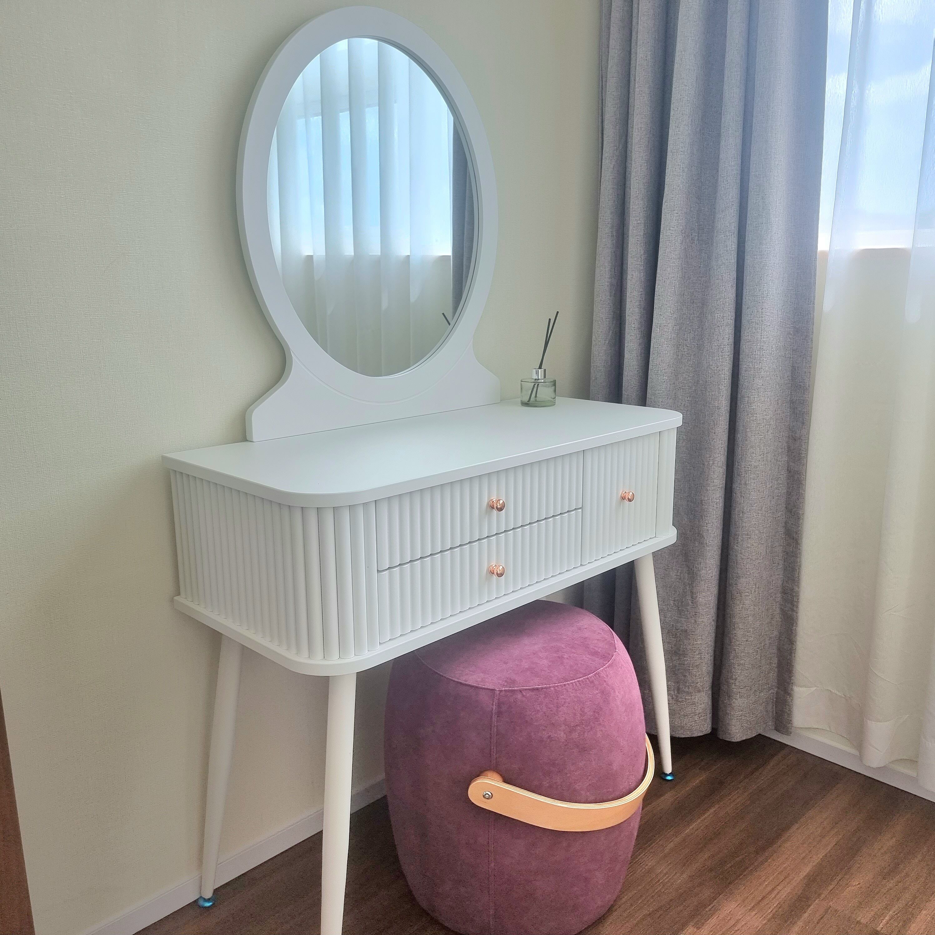 Une coiffeuse blanche avec un miroir, des tiroirs et un pouf violet.