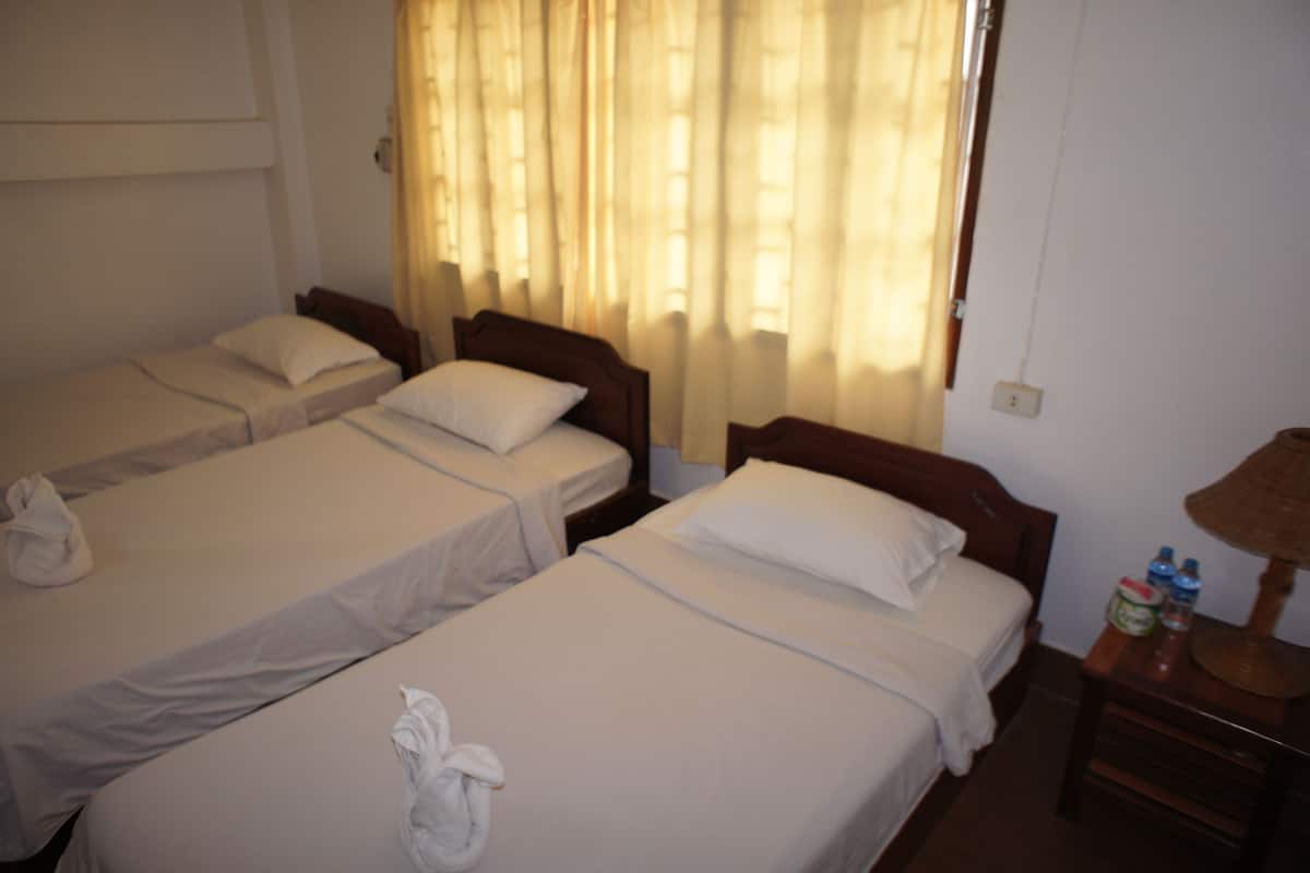 A hotel room with three single beds, each with a white bedspread and pillows. There is a wooden nightstand with a lamp and bottled water.