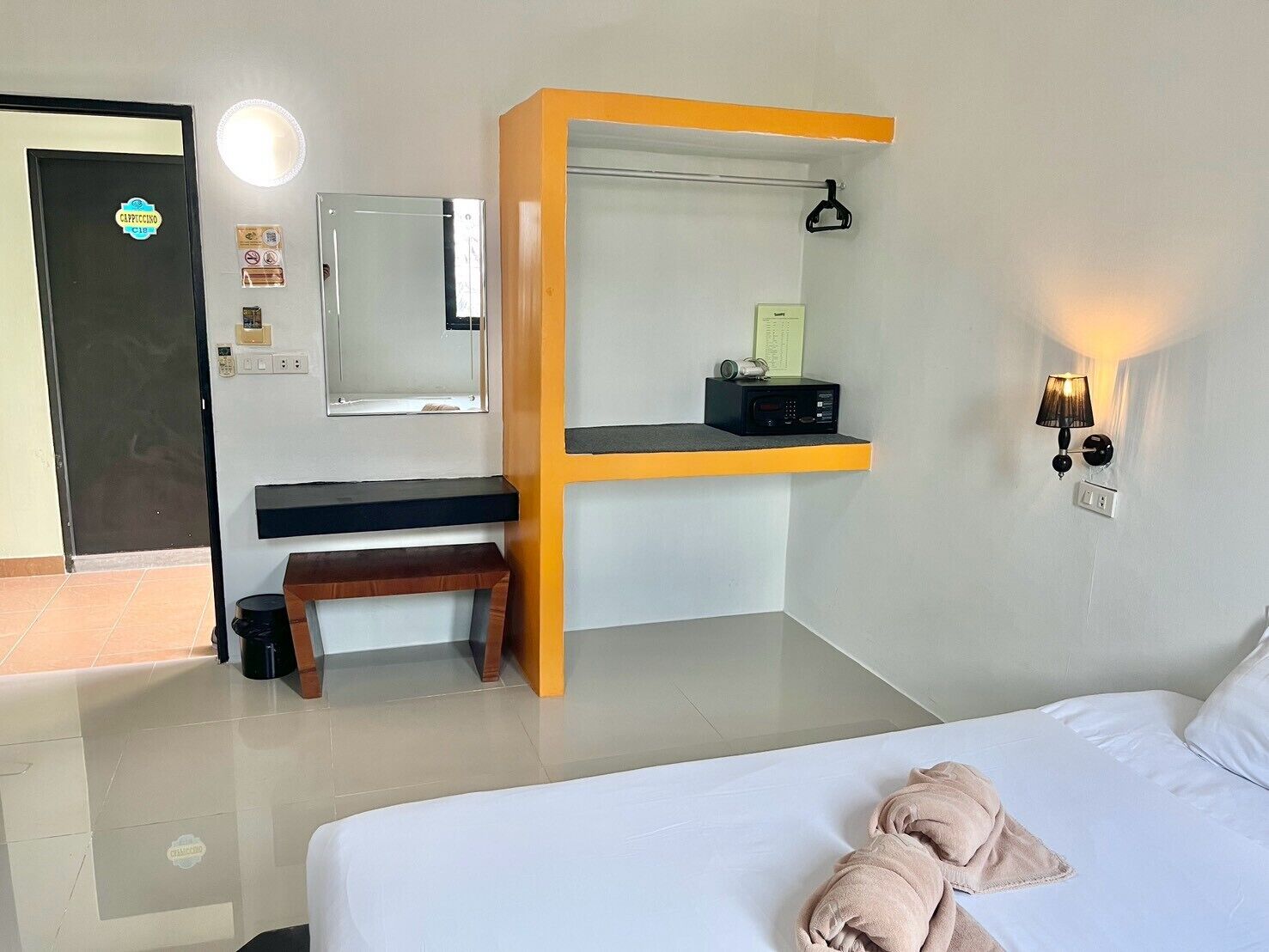 A hotel room with a bed, a desk with a chair, a mirror, a wall-mounted lamp, and a door with a sign.