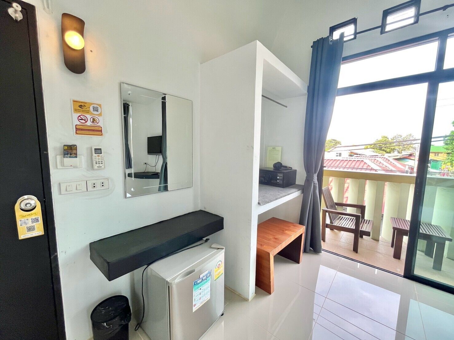 Habitación de hotel con balcón, refrigerador, espejo, estante y ventana con cortinas.