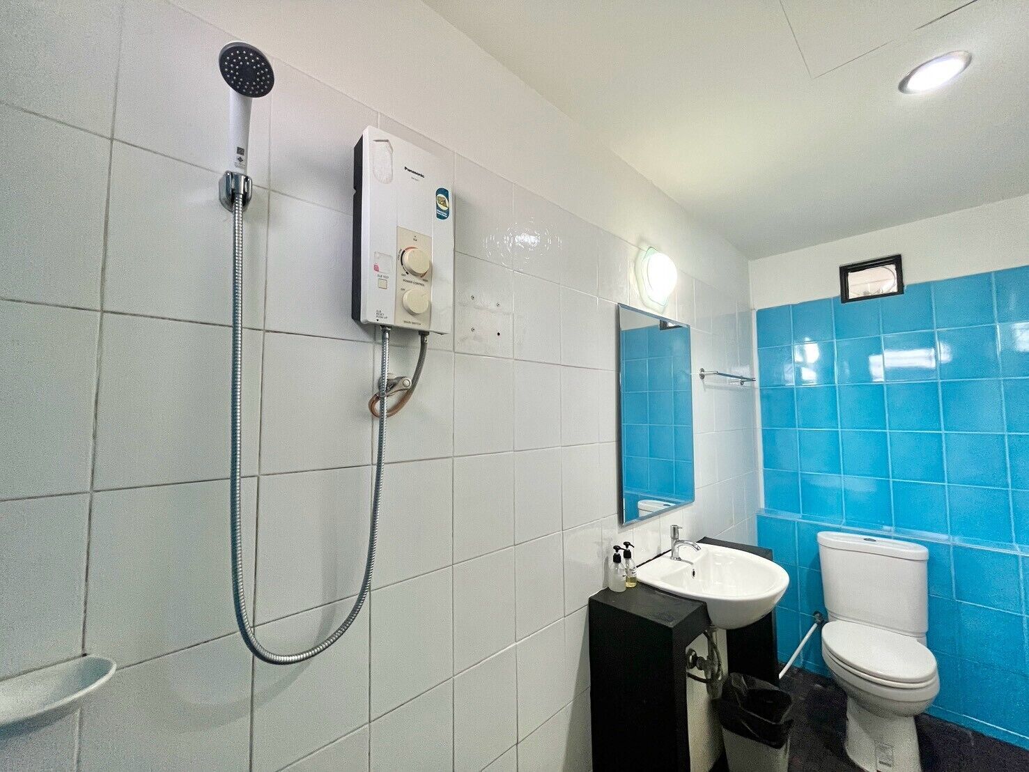 Un baño con paredes de azulejos blancos, zona de ducha con azulejos azules, lavamanos blanco y un inodoro.