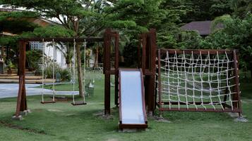 Children can play on the swing set, slide, and rope net in this outdoor play area surrounded by lush greenery and a nearby building.
