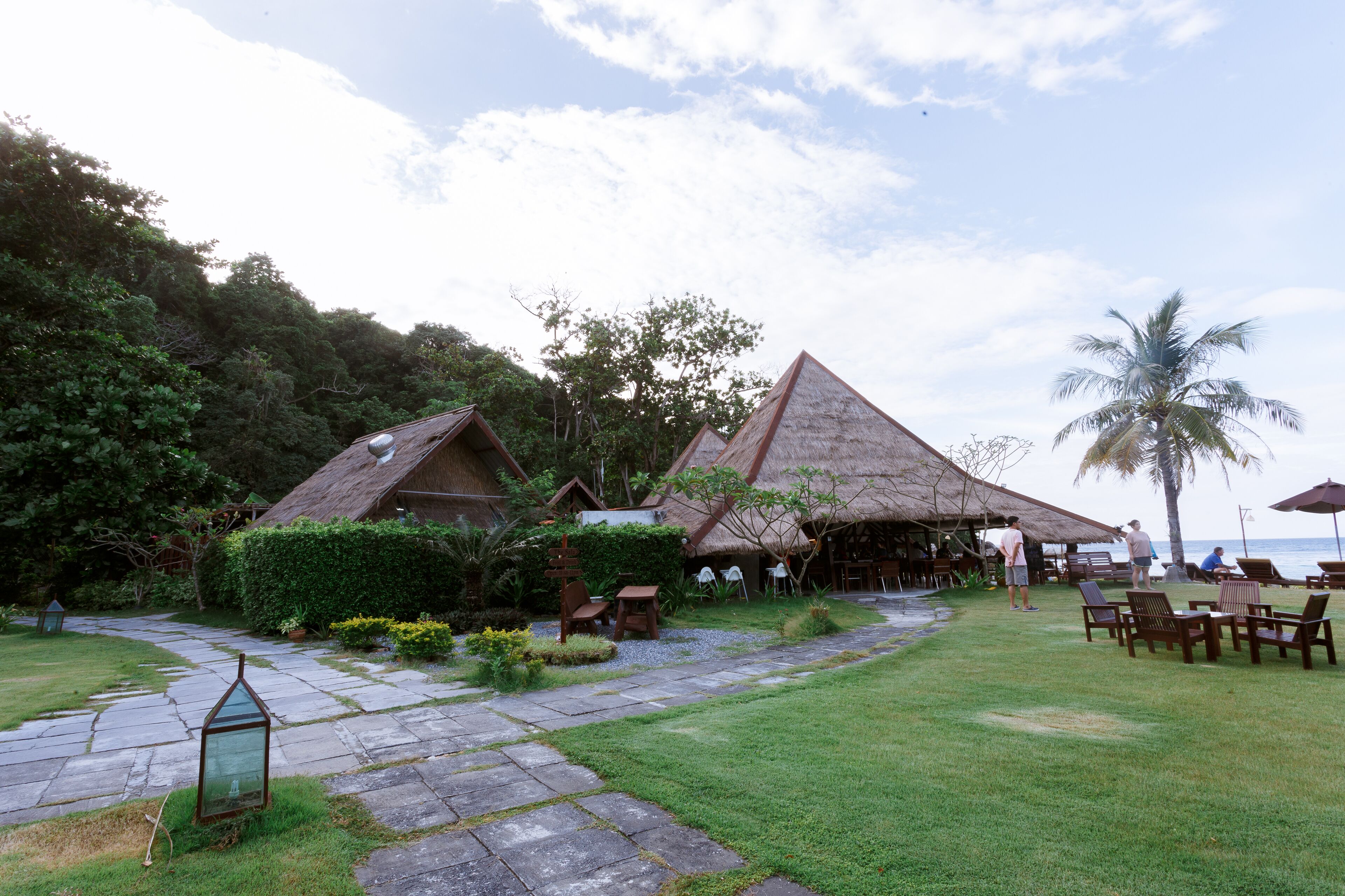 A tropical resort with thatched-roof huts, a paved pathway, and outdoor seating areas.