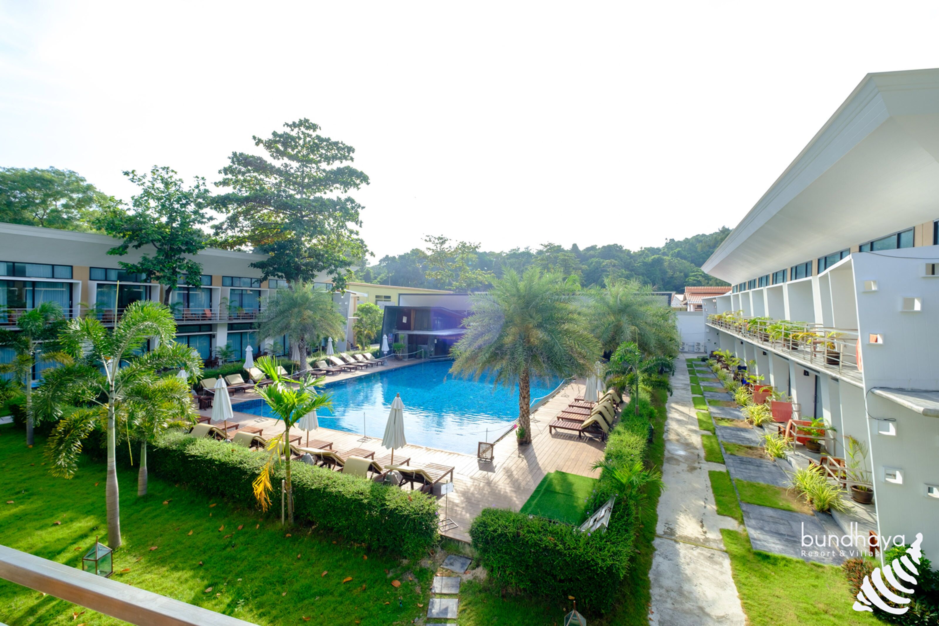 A resort with a swimming pool surrounded by lounge chairs and umbrellas, a well-maintained lawn, and a modern building structure.