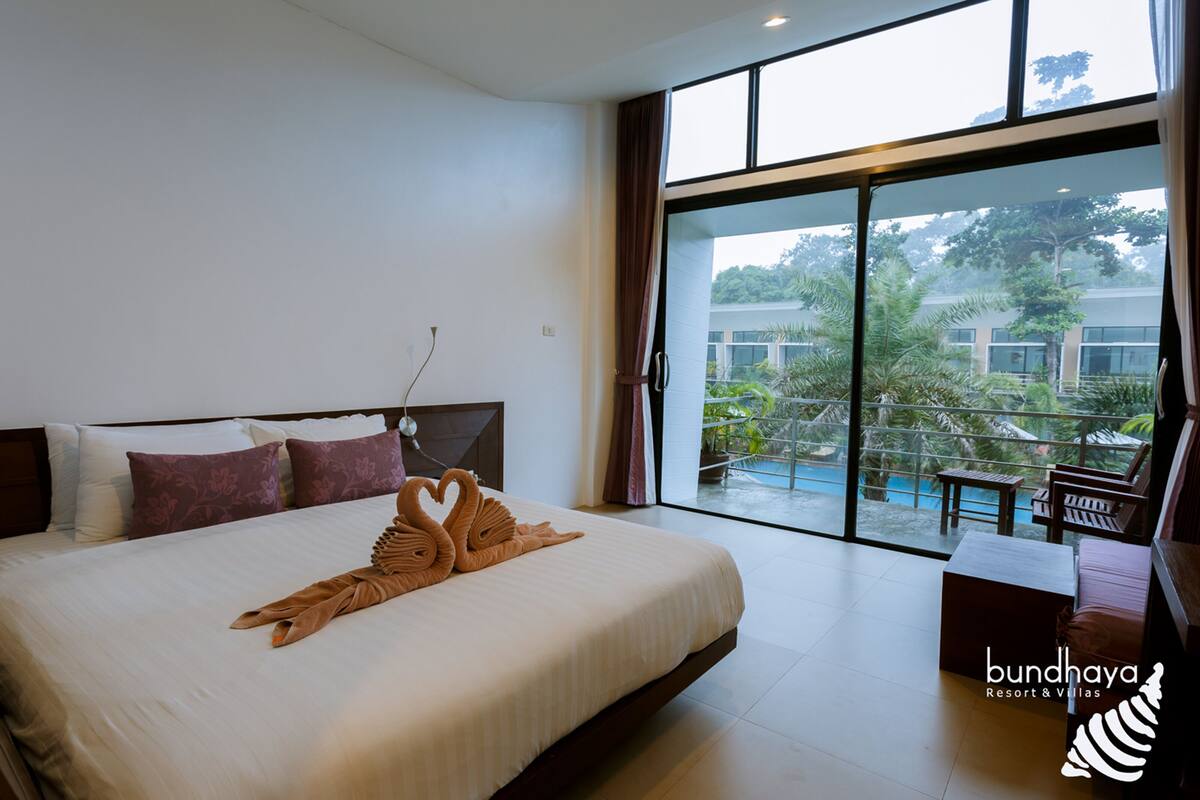 Een hotelkamer met een groot bed, een balkon met uitzicht op een zwembad en groen, en een bordje met 'bundhaya resort & villas'.