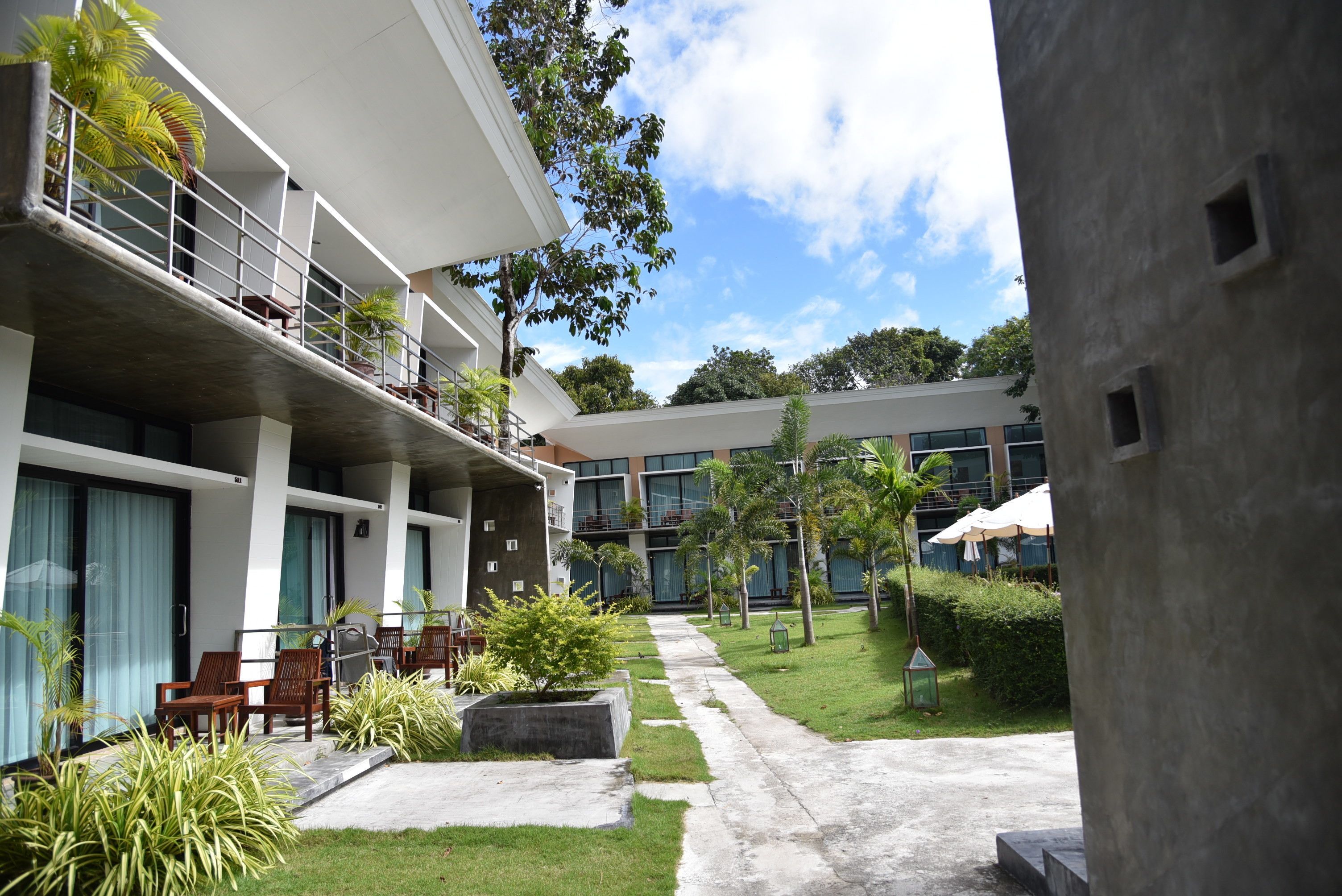A modern hotel with a covered outdoor seating area, a concrete pathway, and lush greenery.