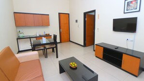 A compact living space with a sofa, a coffee table with fruit, a dining table with chairs, a kitchenette, a TV, and a door leading to another room. - The Star Hill Hotel (Balikpapan)