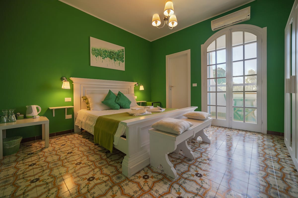 Une chambre avec un mur vert, un lit blanc avec des oreillers, une table de chevet avec une lampe et une fenêtre donnant sur un beau paysage.