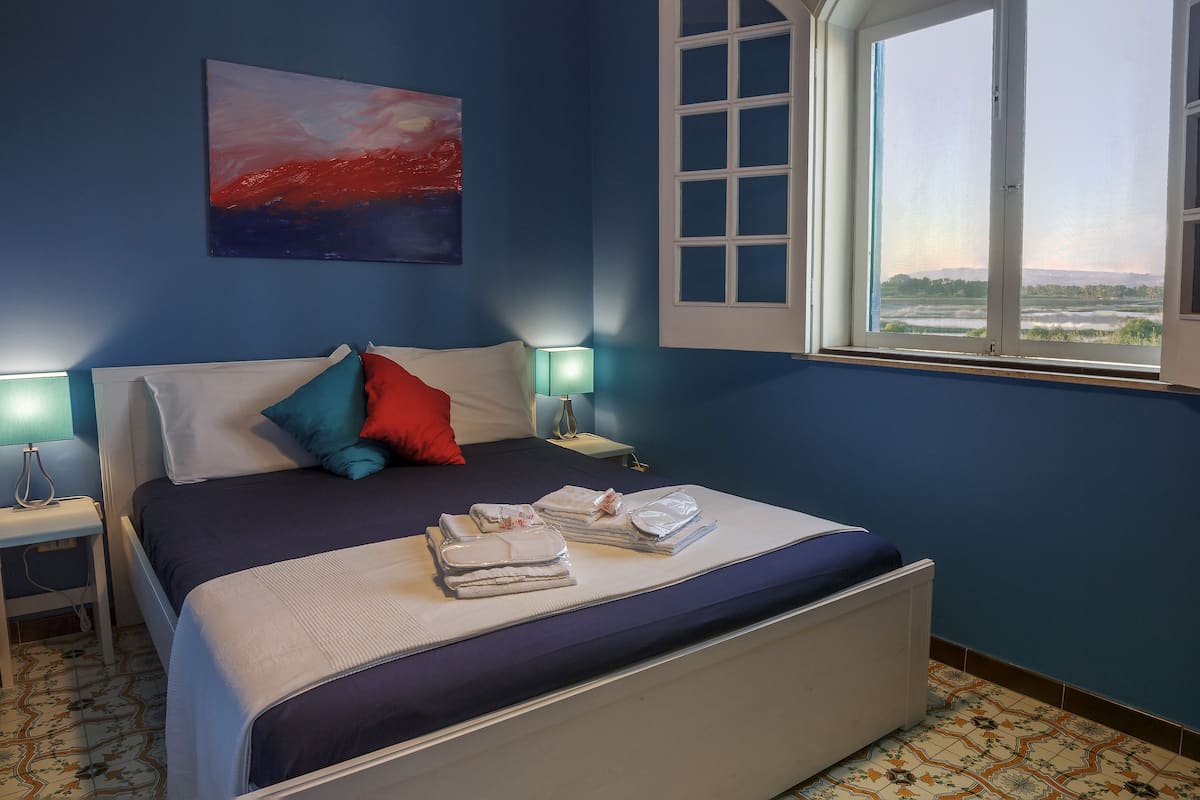 Une chambre à coucher avec un lit, des oreillers, une table de chevet, une lampe, une fenêtre donnant sur un beau paysage et un tableau accroché au mur.