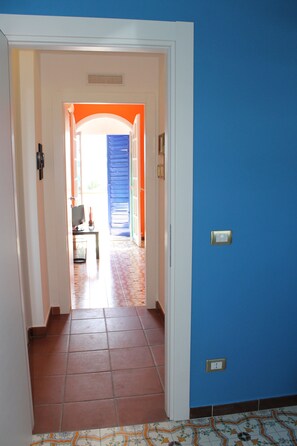Um corredor com parede azul, chão de azulejos e porta que dá para um quarto com televisão e uma mesinha.