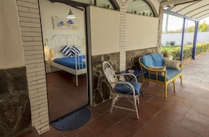 Quarto com cama, duas cadeiras, uma mesinha e vista para um jardim.