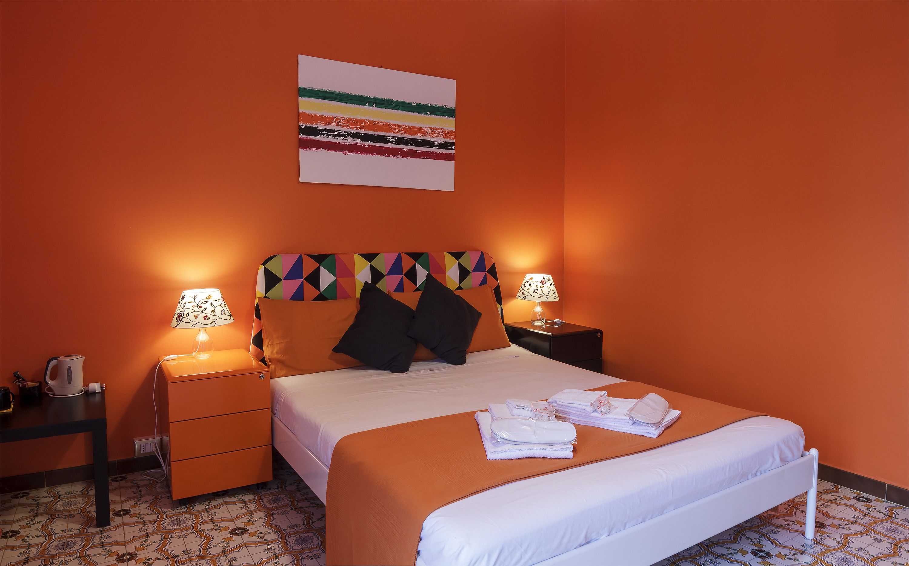 A hotel room with a bed, two bedside tables, a nightstand, a lamp, and a colorful headboard.