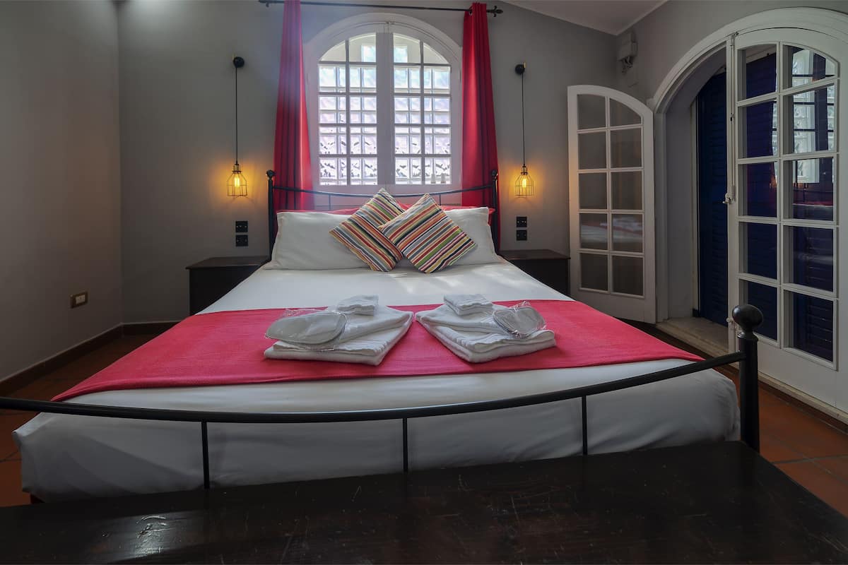 Un lit bien fait, recouvert d’une couverture rouge et agrémenté de deux oreillers, une fenêtre avec des rideaux rouges et une porte à panneaux de verre.