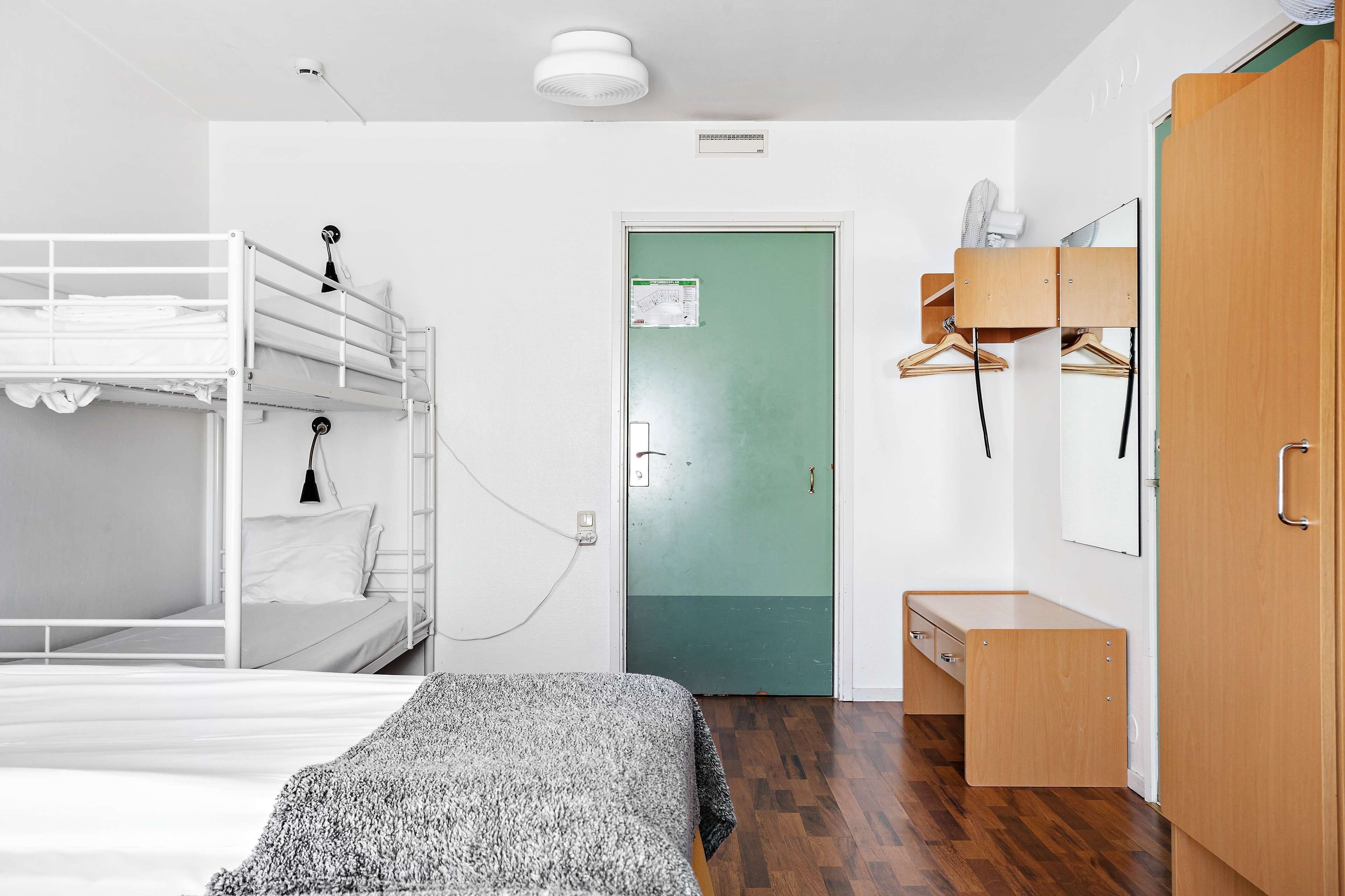 A bunk bed with a bedside table and a small desk with drawers. There is a green door with a white sign, a wooden wardrobe, and a mirror.