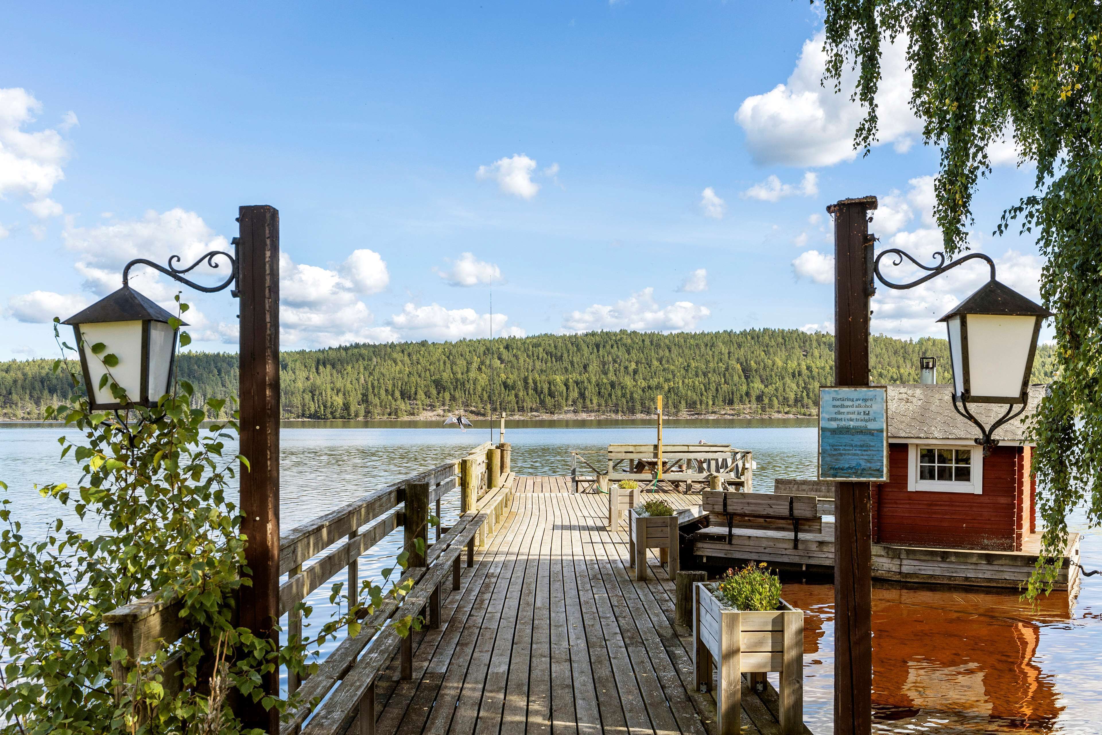 A wooden dock with benches and a small building, overlooking a calm lake with a forested shoreline.