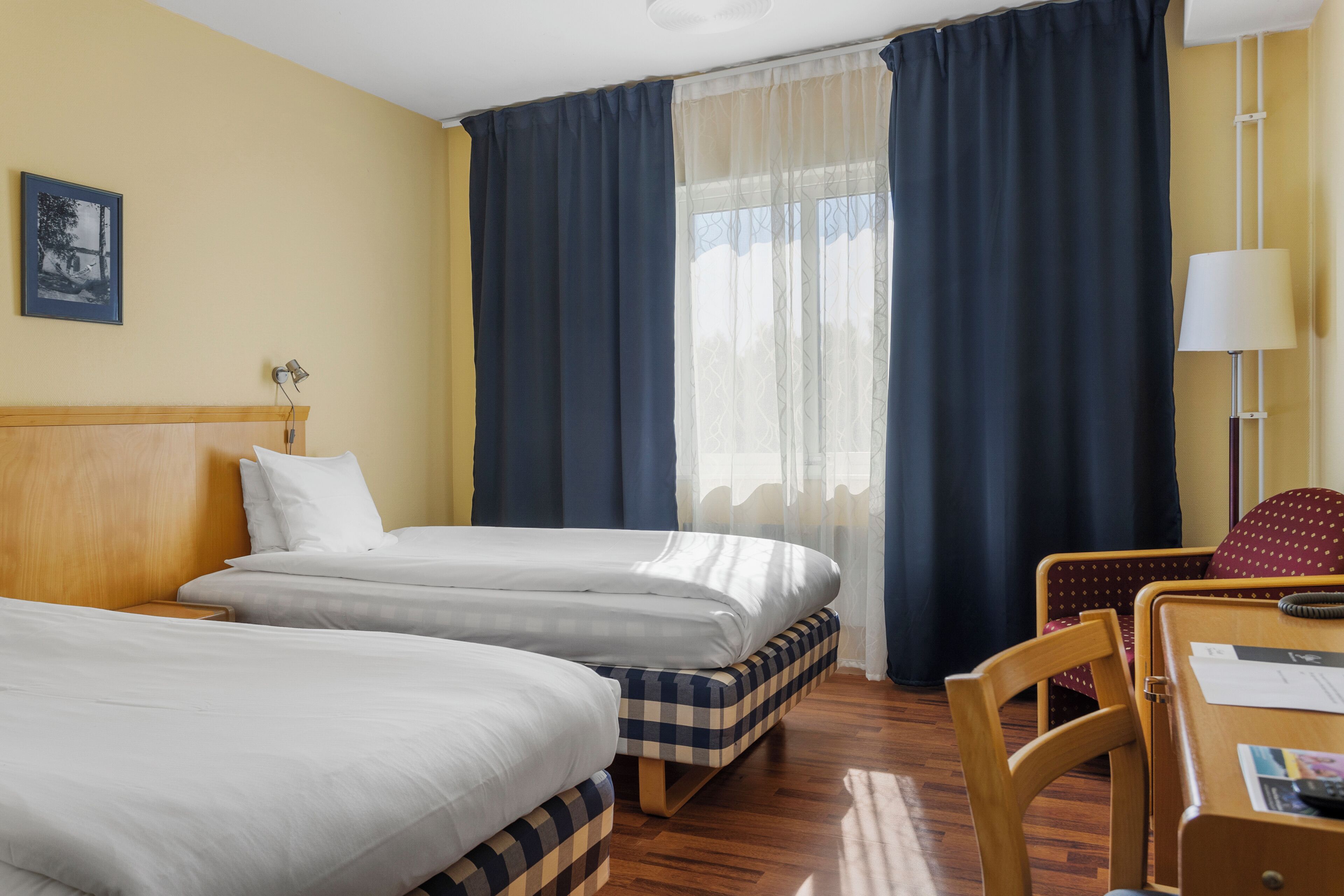 A hotel room with two beds, a desk with a chair, a lamp, and a window with curtains.