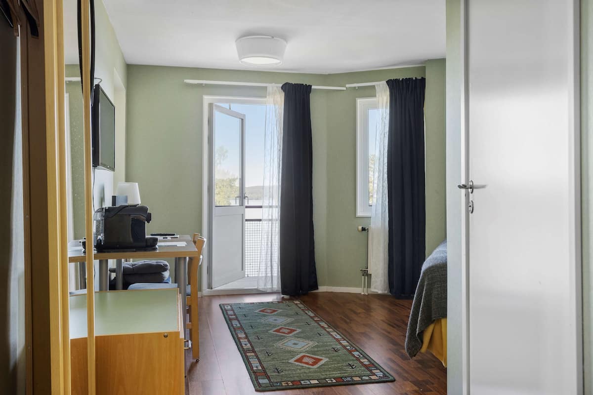 A room with a desk, chair, and a bed. There is a window with curtains, a door, and a view of the outdoors.
